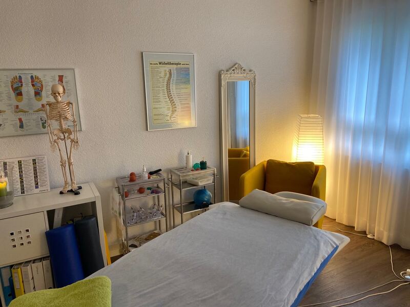 Behandlungsraum mit Therapieliege, gelbem Sessel und anatomischen Wandplakaten.